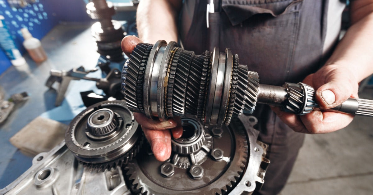 A mechanic holding up a part from a mechanical gearbox from a vehicle. There are multiple helical gears locked together.